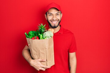 Hispanic man with beard wearing courier uniform with groceries from supermarket looking positive...