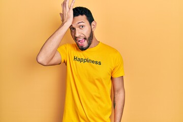 Hispanic man with beard wearing t shirt with happiness word message surprised with hand on head for...