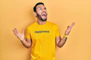 Hispanic man with beard wearing t shirt with happiness word message crazy and mad shouting and yelling with aggressive expression and arms raised. frustration concept.