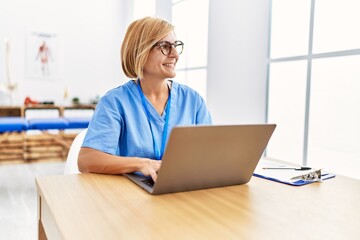 Middle age blonde woman wearing physio therapy uniform using laptop at clinic