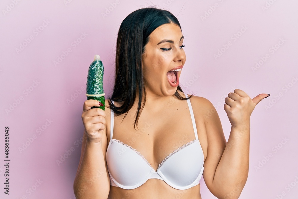 Sticker Beautiful brunette woman holding condom on cucumber for sex education pointing thumb up to the side smiling happy with open mouth