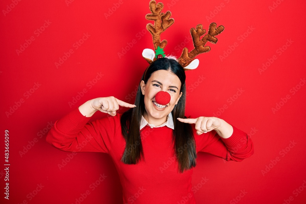 Sticker Young hispanic woman wearing deer christmas hat and red nose smiling cheerful showing and pointing with fingers teeth and mouth. dental health concept.