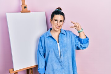 Young hispanic woman standing by painter easel stand smiling and confident gesturing with hand...