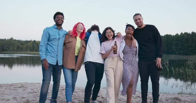 Six people of different nationalities meet each other at the lake, friends spend time together drinking beer and fooling around, embrace each other standing in a row, laugh while leaning forward