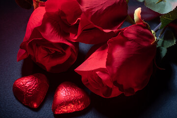 Closeup of heart shaped chocolate confections, red roses and a gift against dark background.