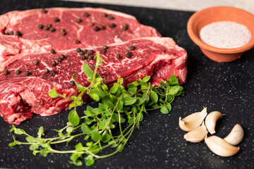 Two raw marbled beef steaks, ready for cooking, with fresh oregano, garlic, and peppercorns.