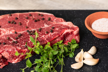 Two raw marbled beef steaks, ready for cooking, with fresh oregano, garlic, and peppercorns.