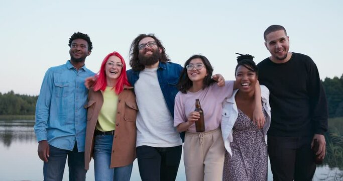 Six people of different nationalities meet each other at the lake, friends spend time together drinking beer and fooling around, embrace each other standing in a row, laugh while leaning forward