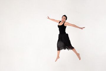 A brunette woman in a black dress barefoot jumped into the air in delight on a white background