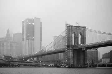 Black and white photo of skyscrapers of Manhattan and Brooklyn bridge on foggy and cloudy day. Famous bridge. Postcard view of New York, USA.