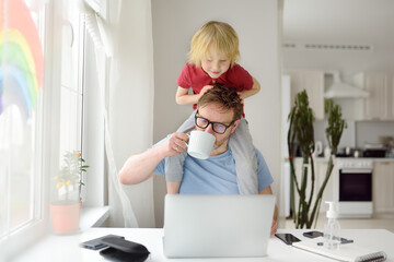 Man is working from home with laptop during quarantine. Home office and parenthood at same time. Exhausted parent with hyperactive child.
