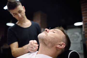 Barber master shaving handsome mature bearded man using straight razor in salon. Hair artist making beard style for person in male barbershop. Services of professional stylist.