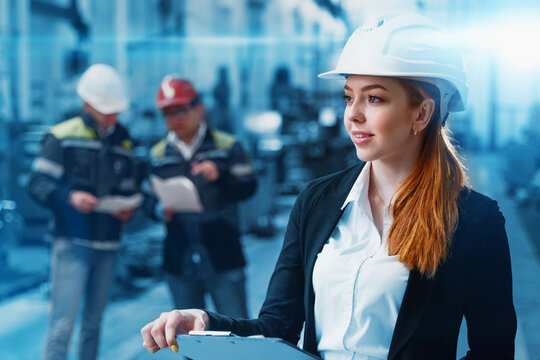 A successful engineer girl checks the technical documentation at the plant. In the background, two workers in helmets and working uniforms.