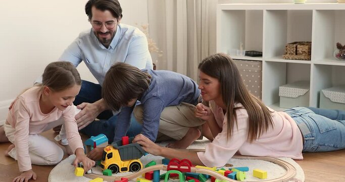 Young couple with little son and daughter spend free time at home, play railroad and wooden cubes lying on warm floor in modern nursery. Concept of family pastime leisure, children development concept