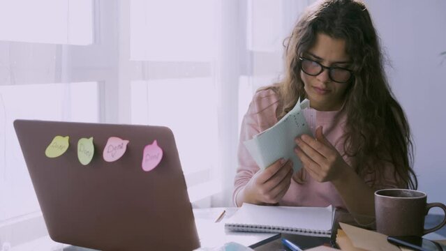 Procrastinated female student with notebooks tired of studying near contemporary laptop at glass table. Overworked woman take off glasses and massage eye during paper work at home