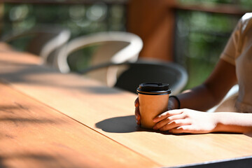 Fototapeta na wymiar Close up with young female adolescence holding take away coffee cup among sunlight while sitting at wooden table.