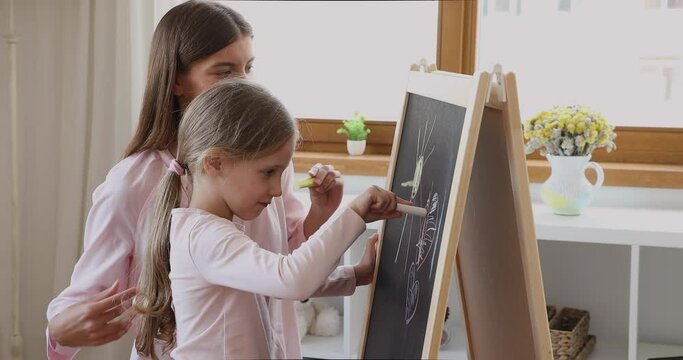 Young loving mother and little cute 6s daughter drawing on board with colored chalks, family having fun enjoy pastime and communication standing in cozy room. Kids development, creative hobby concept