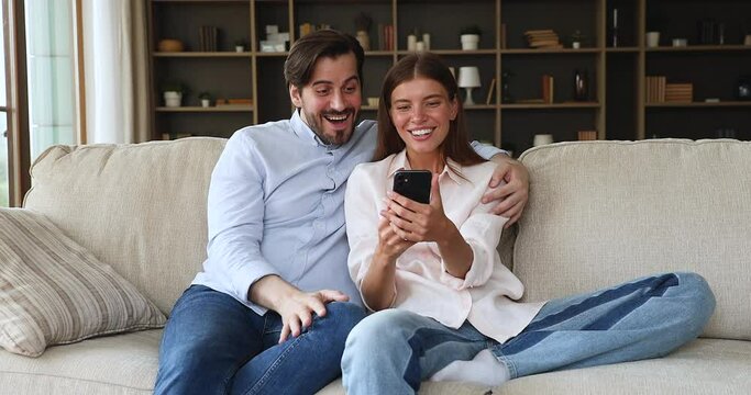 Young 30s couple sit on couch read great news on smart phone, got unbelievable commercial offer, rejoicing long awaited sale gesturing feel overjoyed, exulting after winning on-line lottery concept