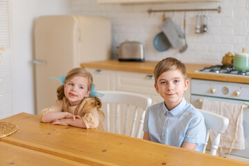 Children sit at table in kitchen and wait for their parents to make them breakfast or lunch. When mom sets the table. Happy time and childhood of caucasian school children