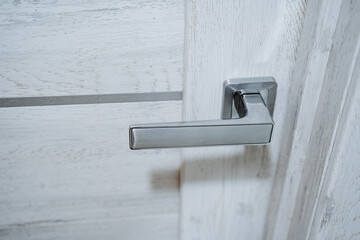 Silver handle at the wooden door. The entrance to the room. The door is made of light wood. Movable element of the door.