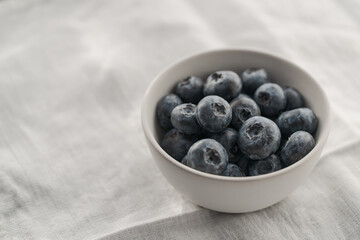 Organic ripe blueberries in white ceramic bowl on white linen cloth