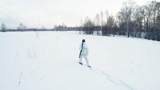 A hunter in winter camouflage and snowshoes walks through a winter forest.