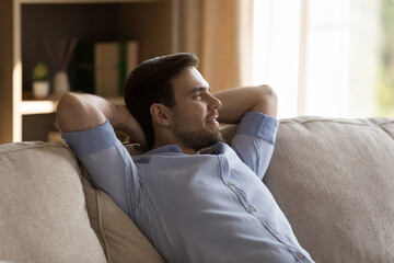 Smiling carefree millennial man resting on comfortable couch with folded hands behind head, looking in distance visualizing future, enjoying peaceful weekend time, breathing fresh air at home.