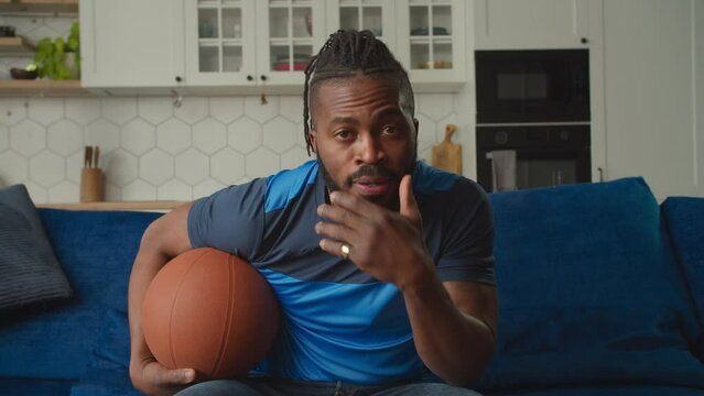 Handsome African American male basketball fan with basketball ball watching match on tv, cheering favorite team and celebrating victory with fist pump gesture, expressing excitement and joy at home.
