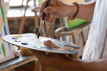 Artist hand holding palette at easel with oil, acrylic paints and paintbrush close up. Art school student learning craft skills, drawing on canvas. Creative hobby, artistic class concept. Cropped shot