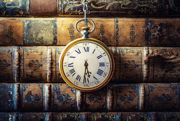 Vintage clock hanging on a chain on the background of old books. Old watch as a symbol of passing time. Concept on the theme of history, nostalgia, old age. Retro style.