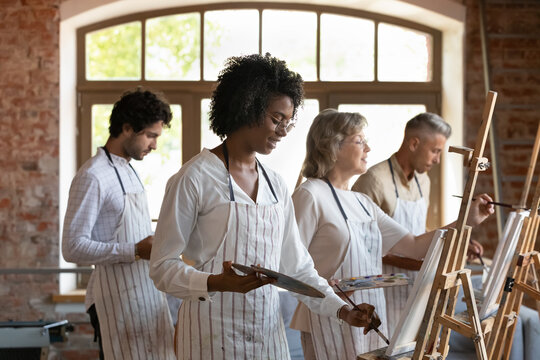 Happy Black art studio student girl in apron drawing at paints at easel, enjoying creative artistic relaxing therapy. Diverse group of different aged artist creating artworks on class