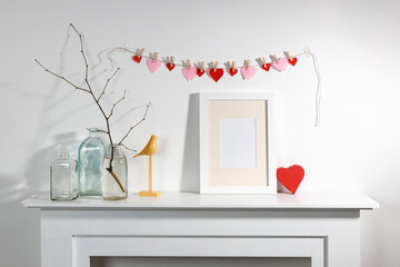 Decoration for Valentine's Day. A garland of felt hearts over a decorative fireplace. Photo frame, a vase with a branch, a gift in a heart-shaped box, a bird figurine on a white shelf panel.
