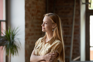 Peaceful thoughtful long haired young woman in casual looking at window away with folded hands, standing in loft space home, thinking, dreaming, smiling at good thoughts, pondering on future plans