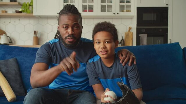 Positive happy black father and lovely school age son with baseball glove and ball sitting on couch, chatting and watching baseball game on tv while enjoying leisure in domestic room during weekend.