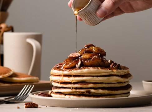 Hand pouring syrup over a plate of maple pecan pancakes with caramelized bananas with coffee set out in the background.