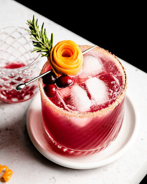 Cranberry orange margarita  on the edge of a counter with an orange peel rose and sprig of rosemary.