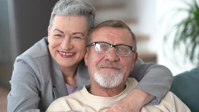 Indore portrait of a gray-haired elderly married couple, a man and a woman hugging and looking at the camera while sitting on the sofa