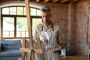 Pensive inspired handsome artist man drawing in paints in artistic studio, standing at easel, thinking over idea for artwork, holding pallet, paintbrush, looking at canvas. Craft hobby, art school