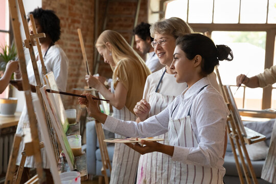 Adult art school Indian student girl in craft apron drawing in paints at easel, holding palette, paintbrush, taking help, advice of mature teacher. Class of artists enjoying creativity in studio