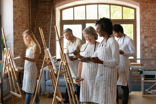 Happy African American art class student in drawing in paints, asking mature mentor for advice, laughing, using pallet, paintbrush at easel. Senior teacher giving help to apprentice with artwork - Powered by Adobe