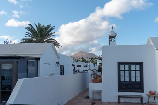 Vista de una calle de un pueblo de Lanzarote con las t&iacute;picas casas blancas de la isla, palmeras y una monta&ntilde;a volc&aacute;nica al fondo en un d&iacute;a soleado, Islas Canarias.