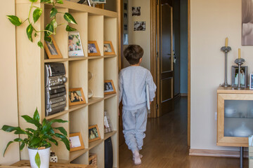 Rear View Of A Small Boy Walking Through The Living Room On His Way To His Bedroom, He Carries A Laptop Under His Arm.