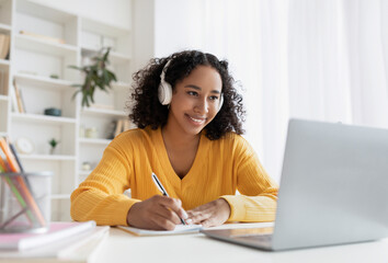 Happy African American woman in headphones having online meeting on laptop, writing down info...