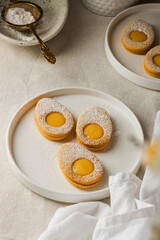 Easter cookies in the form of Easter egg with powdered sugar and lemon cream on a white plate on a light nude concrete background. A greeting card. Easter background. Copy space. Top view