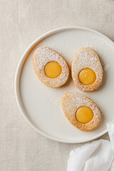 Easter cookies in the form of Easter egg with powdered sugar and lemon cream on a white plate on a light nude concrete background. A greeting card. Easter background. Copy space. Top view