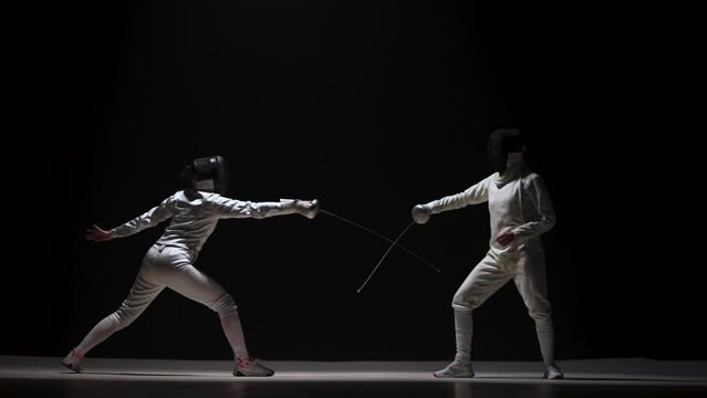Training of female fencers with a rapier on a black background. Athletes in white uniforms and fencing mask practice the skill, attacking the enemy, striking each other, competitive duel. Slow motion.