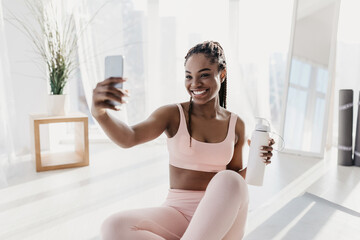 Charming young African American woman with bottle of water taking selfie during home workout