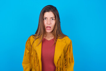 young brunette woman wearing yellow fringed jacket over blue background having stunned and shocked...