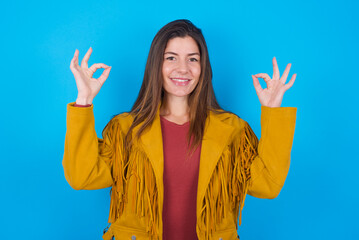 Glad young brunette woman wearing yellow fringed jacket over blue background shows ok sign with...
