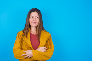 young brunette woman wearing yellow fringed jacket over blue background with thoughtful expression,...